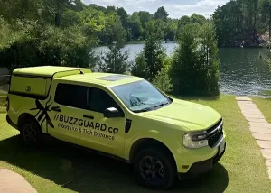 Lime-green BuzzGuard.ca pest control pickup truck parked on a grassy lakeside property with trees and water in the background.